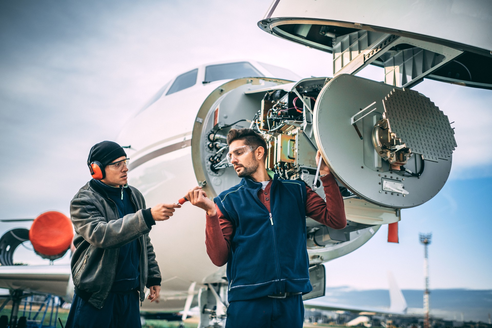 Two aircraft mechanics working on a radar array under the opened up nose cone of a private airplane parked on an airport field
